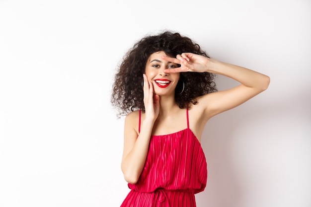 Beleza e moda. Mulher romântica com cabelo encaracolado e vestido vermelho, mostrando o sinal-v e sorrindo feliz para a câmera, em pé sobre fundo branco.