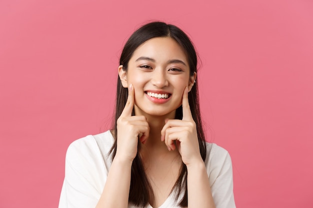 Beleza, emoções das pessoas e conceito de lazer de verão. Close-up de mulher asiática engraçada e bonita com covinhas kawaii, tocando as bochechas e sorrindo feliz, em pé fundo rosa.