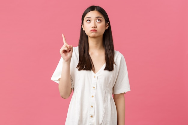 Beleza, emoções das pessoas e conceito de lazer de verão. menina asiática fofa sombria apontando e olhando para o céu, decepcionada com o mau tempo, parecendo chateada com o fundo rosa de pé.