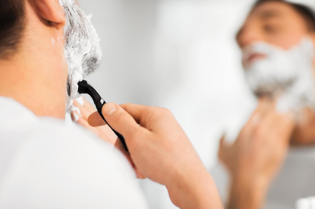 beleza, higiene, barbear, aliciamento e conceito de pessoas - close-up do jovem olhando para o espelho e barba de barbear com lâmina de barbear manual no banheiro de casa