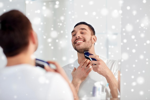 beleza, higiene, barbear, aliciamento e conceito de pessoas - jovem olhando para espelhar e barbear barba com aparador ou barbeador elétrico no banheiro de casa sobre a neve