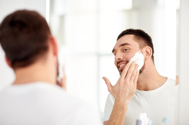 beleza, higiene, barbear, aliciamento e conceito de pessoas - jovem sorridente olhando para o espelho e aplicando espuma de barbear no rosto no banheiro de casa