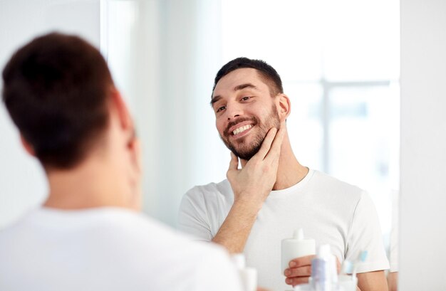 beleza, higiene, barbear, aliciamento e conceito de pessoas - jovem sorridente olhando para o espelho e aplicando loção pós-barba no banheiro de casa