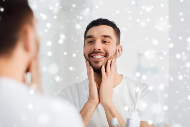 beleza, higiene, barbear e conceito de pessoas - jovem sorridente olhando para o espelho no banheiro de casa sobre a neve