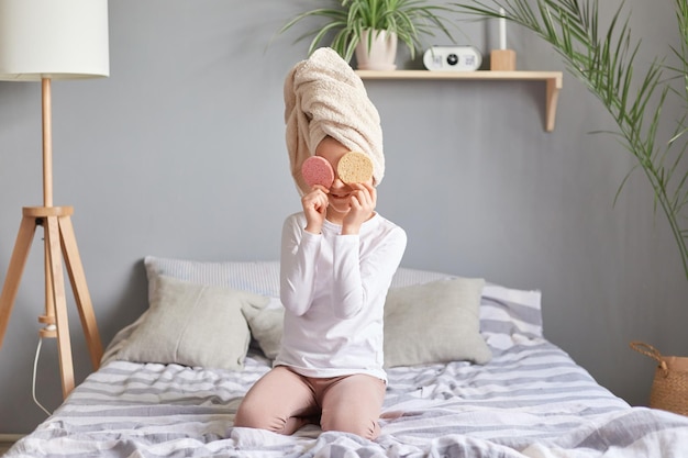 Beleza infantil Procedimentos cosméticos infantis Menina engraçada com camisa branca e toalha na cabeça cobrindo os olhos com esponjas se divertindo após o banho matinal com as ferramentas de cuidados com a pele da mãe