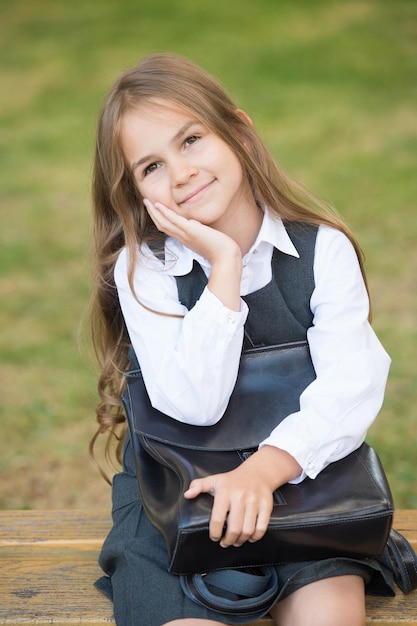 Beleza inteligente. Sorriso de criança feliz sentado no banco. Menina segurar o saco vestindo uniforme. Moda escolar. Salão de beleza. De volta à escola. Educação formal. 1 de setembro. Dia do conhecimento.