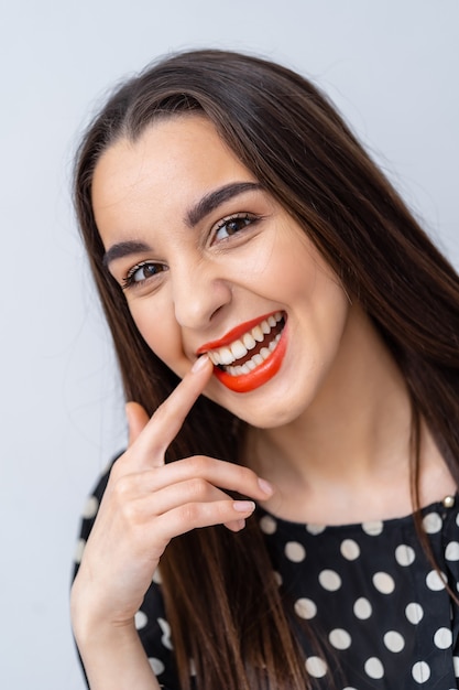 Beleza jovem com sorriso perfeito, lábios vermelhos e dentes brancos. Menina segurando o dedo perto dos lábios vermelhos. Fundo branco.