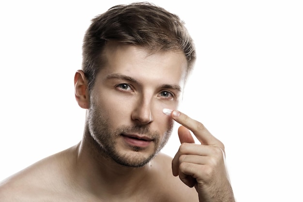 Beleza masculina. Jovem está aplicando um creme hidratante e anti-envelhecimento no rosto contra um fundo branco
