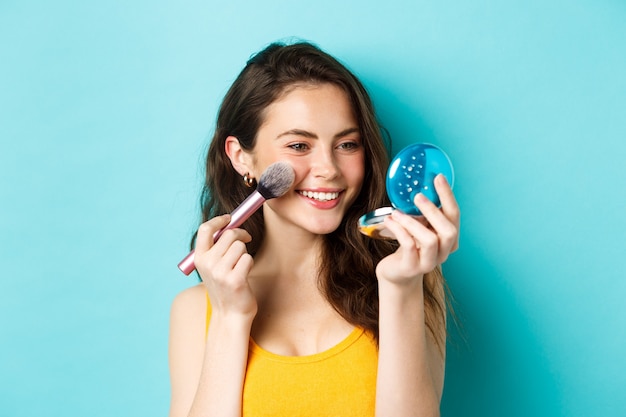 Beleza. Menina moderna com sorriso sincero, aplicando maquiagem com pincel, olhando no espelho de bolso, em pé sobre um fundo azul