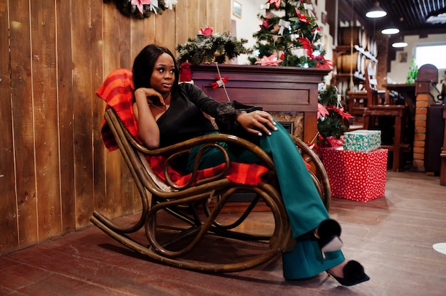 Beleza slim modelo afro-americano usar blusa preta e calças verdes de pernas longas posar contra decorações de Natal com grinalda, lareira e cadeira de balanço.