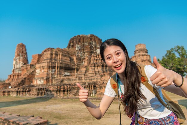 beleza sorridente turista feminina visitando Ayutthaya famoso wat mahathat e mostrando o polegar para cima gesto tirando foto em viagens de Tailândia.