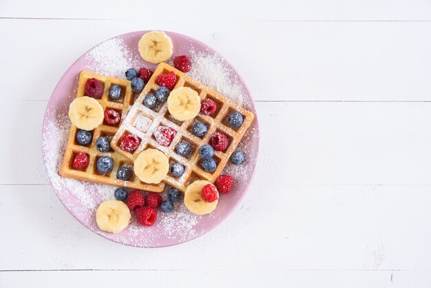 Belgische Waffeln mit Blaubeeren, Himbeeren, Bananen und Zuckerpulver auf weißem Hintergrund. Konzept von leckerem und gesundem Essen. Draufsicht.