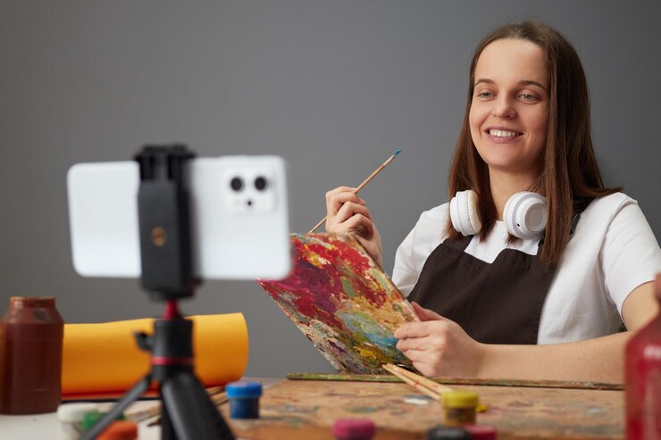 Foto bella atractiva mujer de cabello marrón con delantal marrón pintando usando teléfono móvil en trípode en su lugar de trabajo disfrutando de transmisión en vivo y comunicándose con sus seguidores