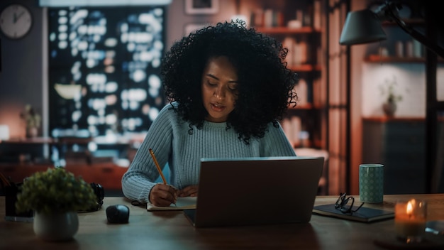 Bella y auténtica mujer latina sentada en un escritorio en una acogedora sala de estar oscura por la noche y usando una computadora portátil en casa Estudiante estudiando en casa Chateando con amigos en redes sociales