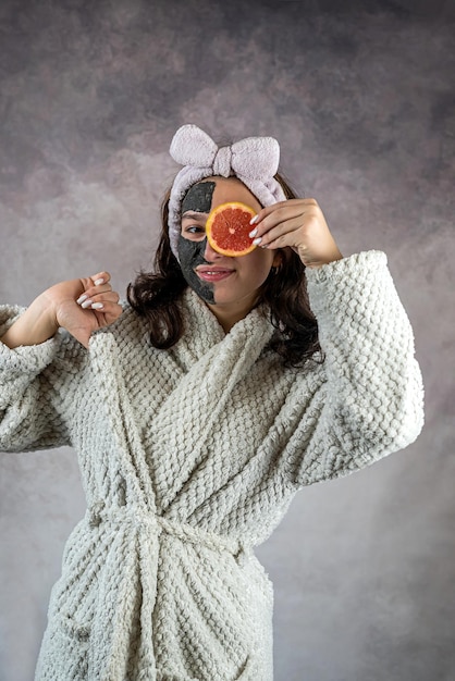 Bella chica en bata blanca con máscara facial negra sosteniendo pomelo aislado procedimientos de spa de cuidado de la piel