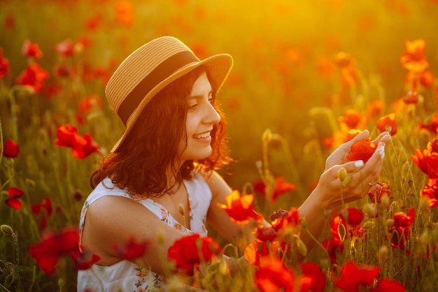 Bella chica en el campo de amapolas al atardecer con un vestido blanco y sombrero