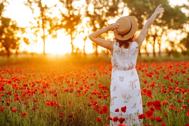 Bella chica en el campo de amapolas al atardecer con un vestido blanco y sombrero