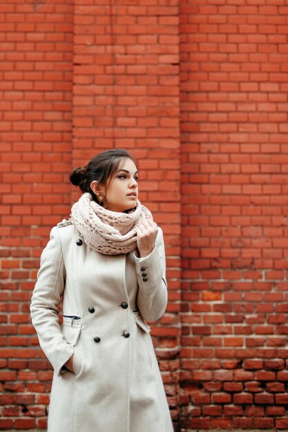 Foto bella chica morena con abrigo blanco y bufanda de pie contra la pared de ladrillo y mirando hacia otro lado