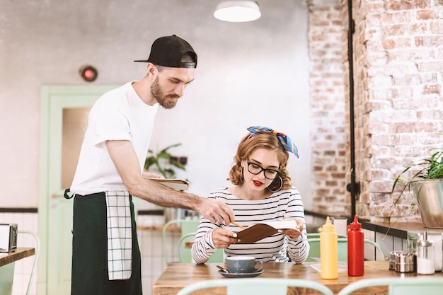 Bella dama con anteojos sentada en la mesa con el menú en las manos y haciendo el pedido mientras el camarero está cerca en el café