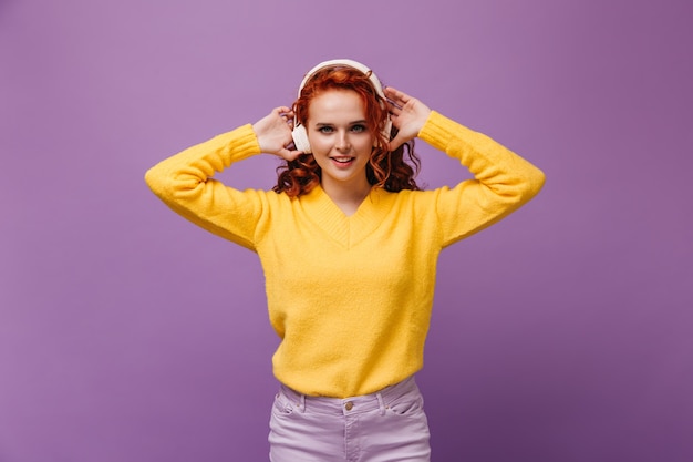 Bella dama en suéter amarillo y auriculares posando sobre pared lila