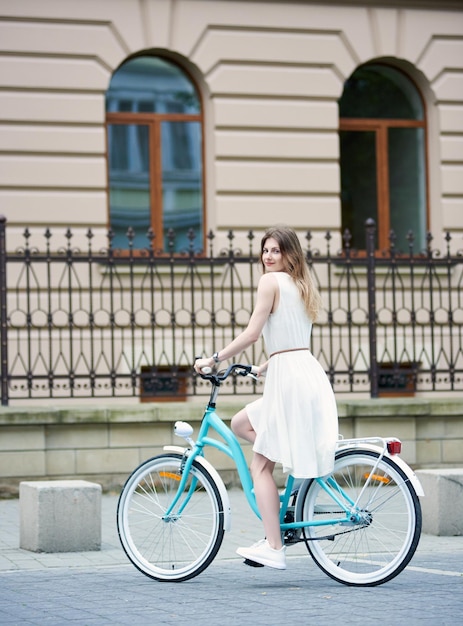 Bella dama en vestido de verano montando bicicleta azul retro en una calle de la ciudad y mirando a la cámara
