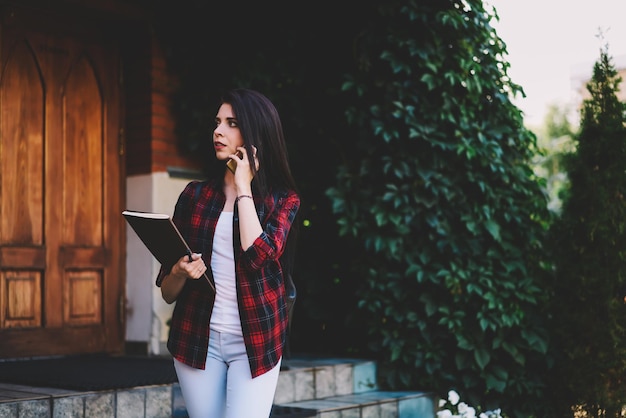 bella estudiante hablando por teléfono inteligente parada en la calle y sosteniendo un libro de texto en la mano