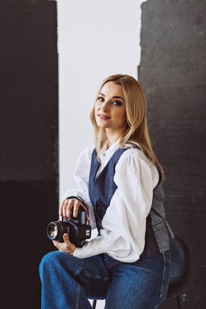 Una bella fotógrafa con un traje casual de mezclilla y una blusa blanca con mangas voluminosas con una cámara en sus manos. Aficiones. Enfoque selectivo suave.