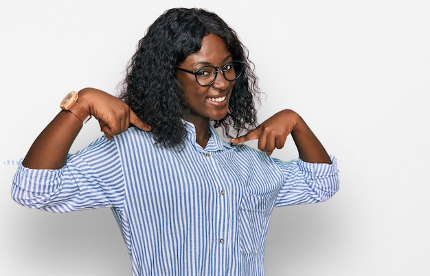 Bella joven africana con ropa casual y gafas mirando con confianza con una sonrisa en la cara señalándose con los dedos orgullosa y feliz