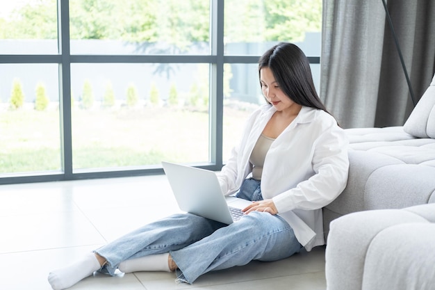 bella joven asiática con pantalones blanco usando una laptop sentada en el suelo en un interior moderno y luminoso