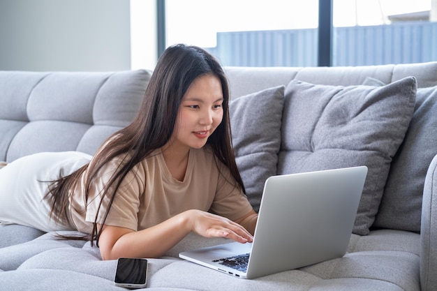 bella joven asiática con ropa informal y acogedora usando una laptop sentada en un sofá en un apartamento moderno