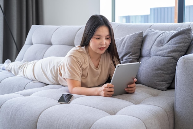 bella joven asiática con ropa informal y acogedora usando una tableta sentada en un sofá en un apartamento moderno
