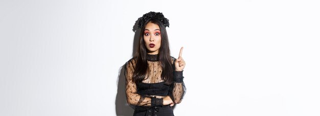 Foto bella joven asiática en vestido de encaje negro y maquillaje gótico han pensado chica en disfraz de bruja levantando el dedo sugiriendo persona idea de halloween