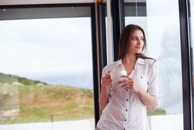 bella joven bebe el primer café de la mañana en el moderno interior de la casa con gotas de lluvia en el cristal de la puerta de la ventana grande