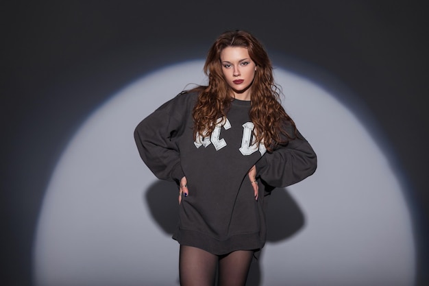 Bella joven elegante con un peinado en una sudadera de moda con medias posando en el estudio sobre un fondo oscuro