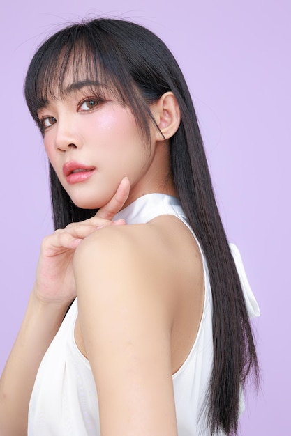 Bella joven modelo asiática de cabello largo con maquillaje natural en la cara piel limpia en fondo violeta aislado retrato de chica linda tratamiento facial cuidado corporal belleza y spa