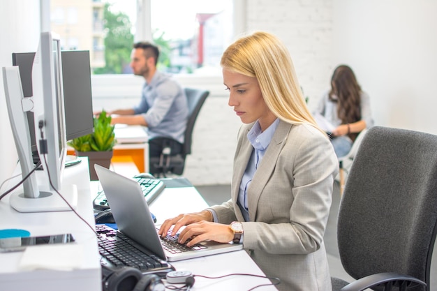 Bella joven rubia con ropa formal trabajando en una computadora portátil en su lugar de trabajo