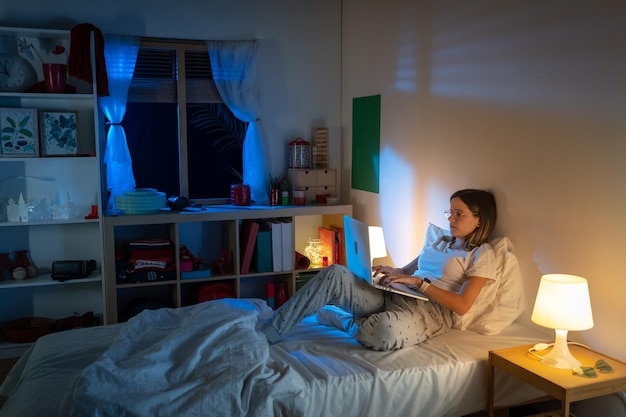 Bella joven sentada en la cama por la noche trabajando con su portátil en medio de la noche