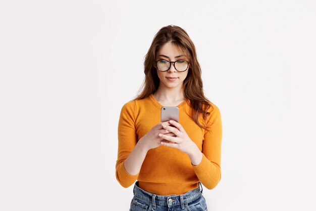 Una bella morena con gafas y un suéter amarillo responde mensajes en su teléfono