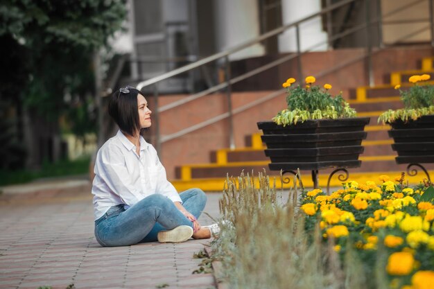 bella morena se sienta en el suelo cerca del lecho de flores de la ciudad con flores amarillas