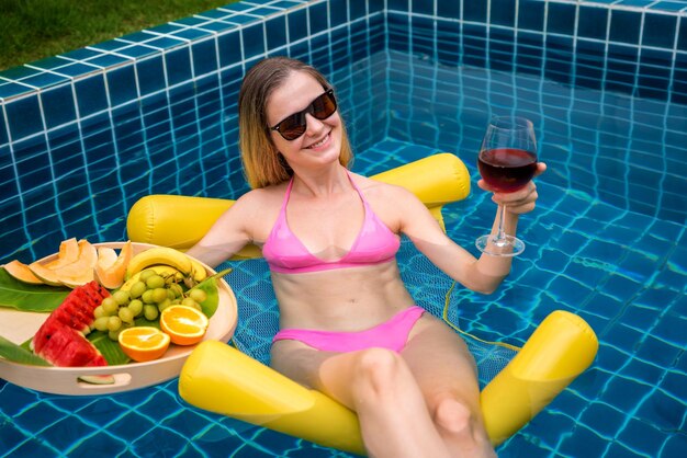 Bella mujer acostada en una hamaca flotante en la piscina con vino y bandeja de frutas