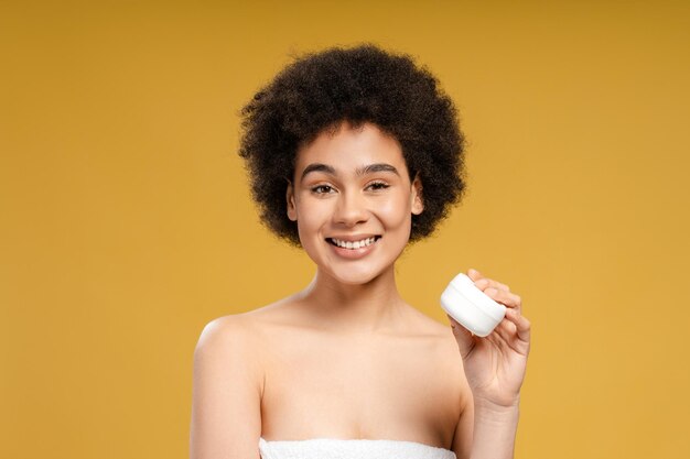 Bella mujer afroamericana con cabello rizado sosteniendo una botella blanca de crema mirando a la cámara