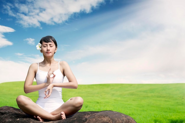 Bella mujer asiática meditando al aire libre en la roca en el prado contra el cielo azul