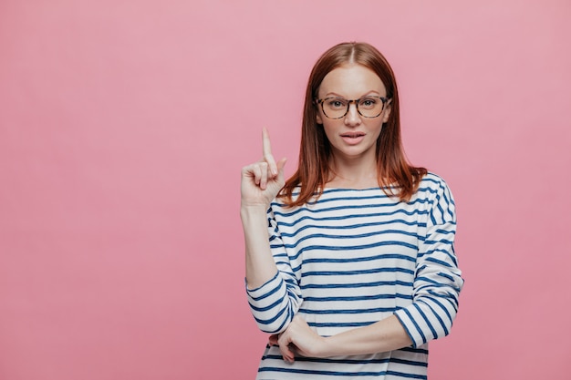 La bella mujer atenta usa gafas y un suéter a rayas, puntos con el dedo índice hacia arriba