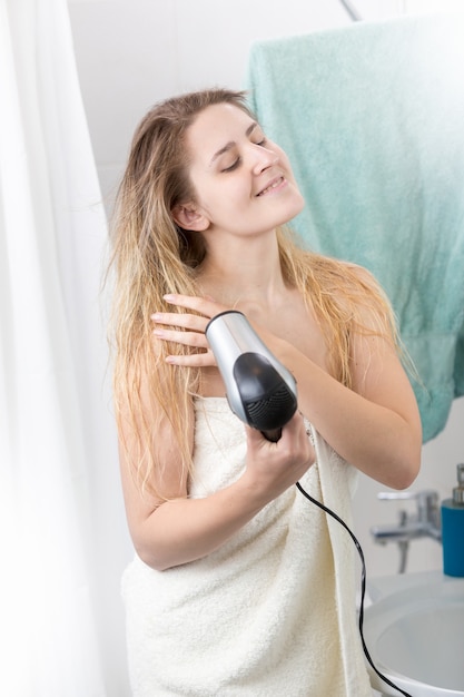 Bella mujer cubierta con una toalla de baño con secador de pelo en el baño.