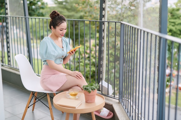 Bella mujer desayunando en el balcón, mientras usa su teléfono