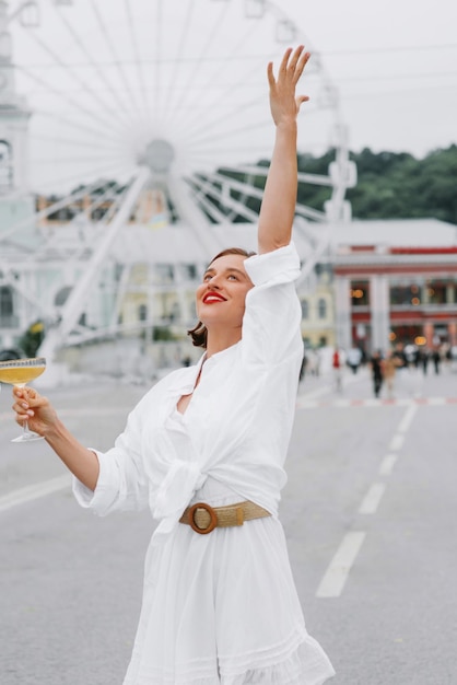 Foto bella mujer feliz es vestido blanco está de pie con la copa de vino en la plaza de la ciudad con el