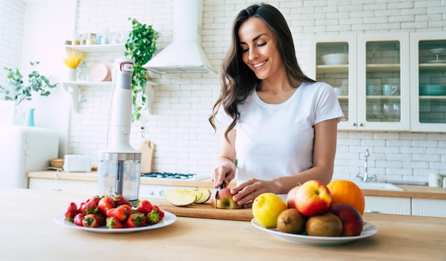 Bella mujer haciendo batidos de frutas con licuadora. Concepto de estilo de vida de alimentación saludable retrato de hermosa mujer joven preparando bebida con plátanos, fresas y kiwi en casa en la cocina