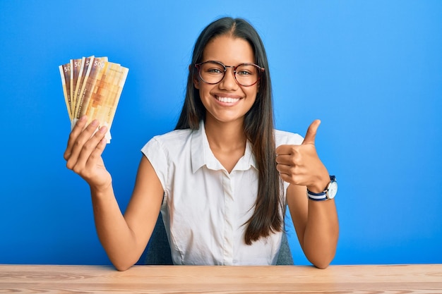 Bella mujer hispana sosteniendo billetes de 500 coronas noruegas sonriendo feliz y positiva pulgar hacia arriba haciendo excelente y señal de aprobación