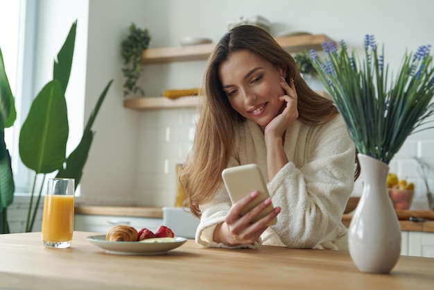 Bella mujer joven en bata de baño usando teléfono inteligente mientras pasa tiempo en la cocina moderna