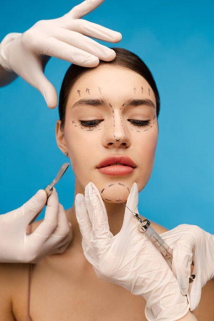 Bella mujer joven lista para una operación antienvejecimiento aislada en fondo azul Médico cirujano plástico haciendo inyección de tratamiento de rejuvenecimiento en el mentón del paciente Cosmetología lifting facial